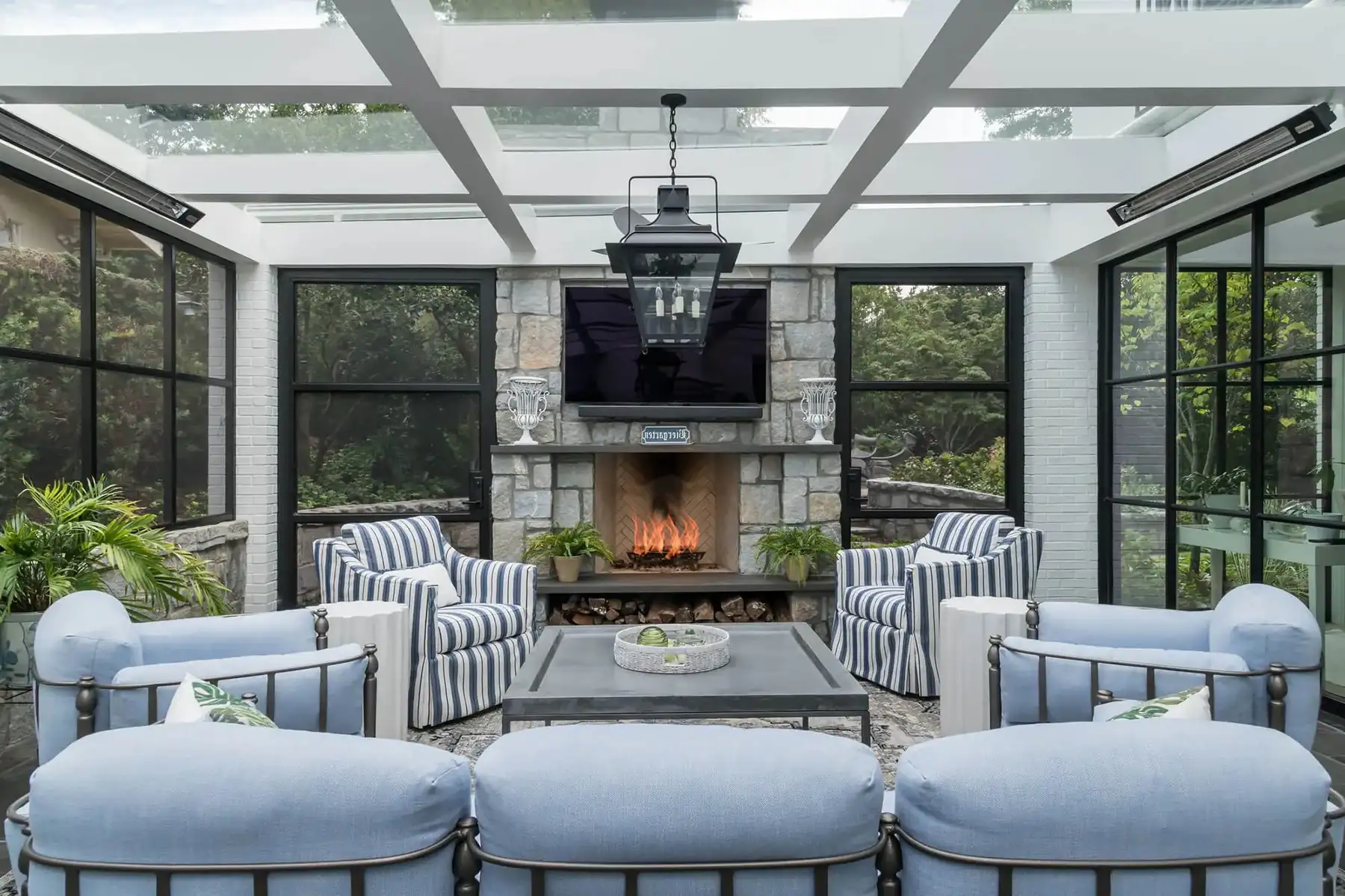 Glass-enclosed sunroom with fireplace, blue lounge seating, and steel-framed windows by The Home X.
