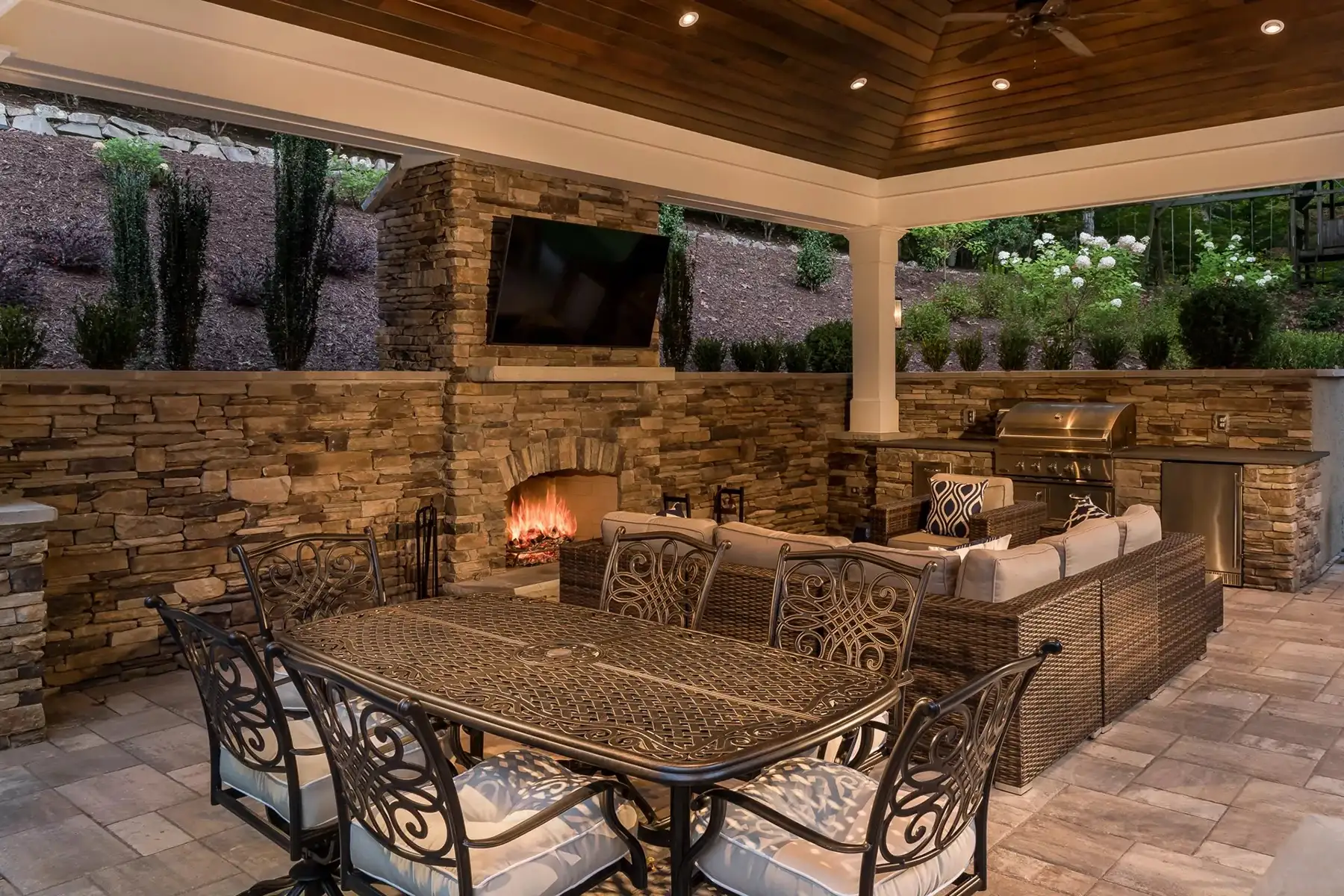 Sergeantsville Rd, Delaware Twp., 08559 11 | The Home X Covered outdoor dining area with rustic stone walls, chandelier lighting, and metal chairs beneath a wood-paneled ceiling.