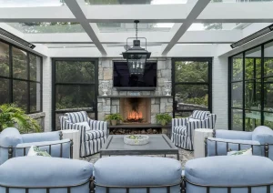 Glass-enclosed sunroom with fireplace, blue lounge seating, and steel-framed windows by The Home X.
