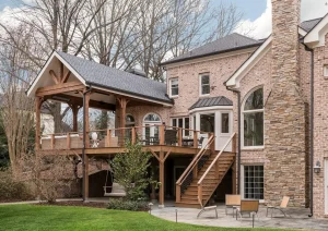 Rear view of a luxury stone home featuring a covered deck, turret-style bay window, and lush wooded surroundings.