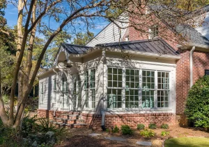 Backyard view of classic white sunroom addition with French doors, brick skirt, and mature landscaping.