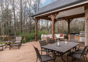 Covered patio with wood beam pergola, outdoor dining set, and stone accents surrounded by a forested backdrop.