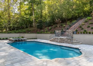 Backyard pool surrounded by stone retaining wall, stairs, and forest backdrop.