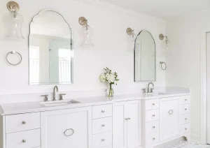 Double sink vanity with arched mirrors, quartz countertops, and brass fixtures in an all-white bathroom.