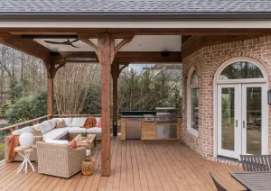 Covered deck with exposed wood beams, paver flooring, arched French doors, and cozy outdoor seating area.