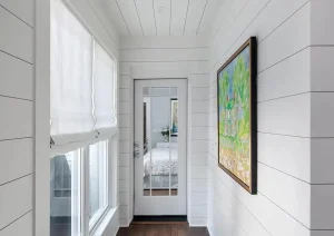 Bright enclosed hallway with shiplap walls, picture windows, and dark wood flooring.