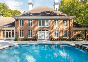 Luxury pool with sun deck and steps, positioned in front of a grand brick home with arched windows and upper balcony.