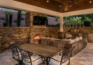Covered outdoor dining area with rustic stone walls, chandelier lighting, and metal chairs beneath a wood-paneled ceiling.