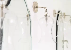 Elegant glass and chrome vanity lights mounted above a double mirror in a high-end white bathroom.