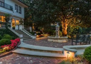 Brick garden steps and fountain illuminated at dusk with landscape lighting and surrounding trees.